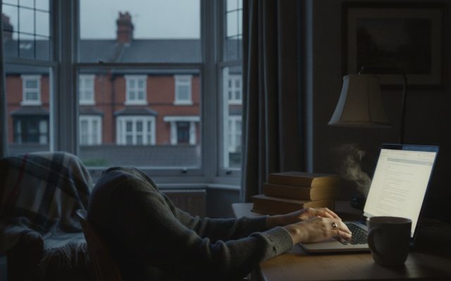 The glow of a laptop screen on a desk in a dim British room at night, hands resting on the keyboard